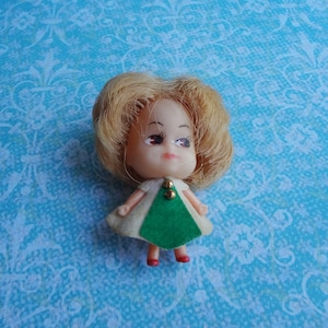 May include: A small plastic doll with blonde hair, wearing a white and green dress. The doll has painted eyes and a painted smile.