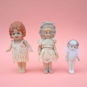 May include: Three antique porcelain dolls on a pink background. The dolls are dressed in vintage clothing, including a fringed dress, a lace dress, and a white outfit with a tulle collar. The dolls have painted faces and detailed features.