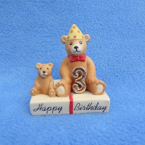 May include: A porcelain figurine of two teddy bears, one wearing a yellow and red party hat, sitting on a white box with the words "Happy Birthday". The larger bear is holding a gold number "3".