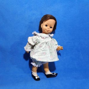 May include: A vintage doll with brown hair, wearing a light green dress and matching bloomers with a floral pattern and white lace trim. The doll has black shoes and white socks. The doll is standing on a blue background.