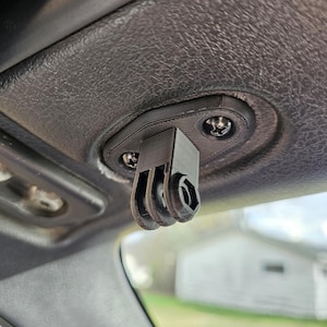Op de afbeelding: Een zwarte, 3D-geprinte camerabevestiging bevestigd in het interieur van een auto. De houder is ontworpen voor een camera, met een drietandige interface en een zeskantige aansluiting. Bevestigd met schroeven aan de hemelbekleding van het voertuig.