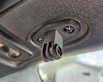 May include: A black 3D-printed camera mount attached to a car's interior. The mount is designed to hold a camera, with a three-prong interface and a hexagonal socket. The mount is secured with screws to the vehicle's headliner.