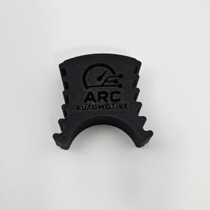Könnte beinhalten: Ein schwarzes Autoteil mit dem Text "ARC AUTOMOTIVE" und einer Tachometergrafik. Das Teil hat eine gebogene Basis und rechteckige Aussparungen. Hergestellt aus mattem, schwarzem Material. Automobil-Design.