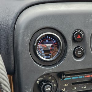 May include: A black gauge with a white and red face, displaying pressure readings in in. Hg and psi. The gauge is labeled "BOOST" and is mounted in a car's dashboard. Other controls and vents are visible.