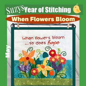 Op de afbeelding: Een kleurrijk geborduurd wandkleed met de tekst "Wanneer bloemen bloeien... bloeit ook de hoop". Het ontwerp bevat een verscheidenheid aan bloemen in tinten rood, oranje, geel, paars en groen. De achtergrond is een lichtblauwe stof met een groene rand.