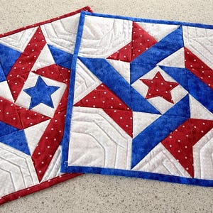 May include: Two square quilted placemats with a patriotic star design. Each mat features a central star in blue or red, surrounded by red polka dot and blue fabric, and white sections. The edges are finished with a coordinating border.