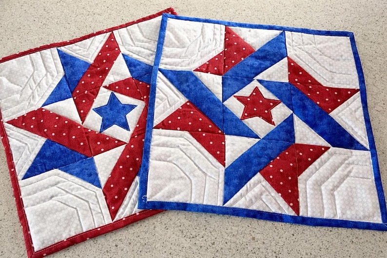May include: Two square quilted placemats with a patriotic star design. Each mat features a central star in blue or red, surrounded by red polka dot and blue fabric, and white sections. The edges are finished with a coordinating border.