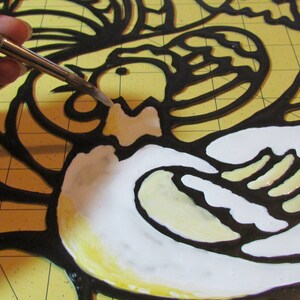 May include: A close-up of a stained glass project in progress. The artist is using a brush to apply white paint to the glass. The glass is outlined in black and has a yellow background.
