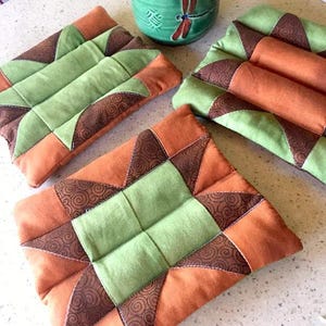 Pode incluir: Quatro bases para copos de tecido acolchoado em tons de verde, laranja e castanho. As bases para copos têm um design geométrico de estrela com um quadrado verde central e detalhes castanhos estampados. Uma caneca verde com um desenho de libélula é visível.