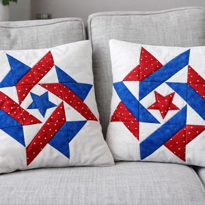 May include: Two white square throw pillows with a patriotic star design. Each pillow features a central star and surrounding starburst pattern in red, white polka dots, and blue. The pillows are on a gray sofa.