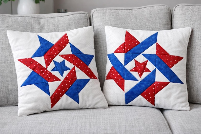 May include: Two white square throw pillows with a patriotic star design. Each pillow features a central star and surrounding starburst pattern in red, white polka dots, and blue. The pillows are on a gray sofa.