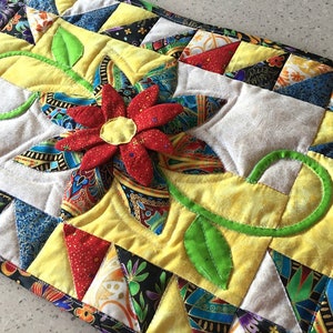 Könnte beinhalten: Ein gesteppter Tischläufer mit einer roten Blumenapplikation und grünen Blättern. Der Läufer ist aus Patchwork-Stoff in verschiedenen Farben und Mustern gefertigt, darunter Gelb, Blau, Rot und Schwarz.
