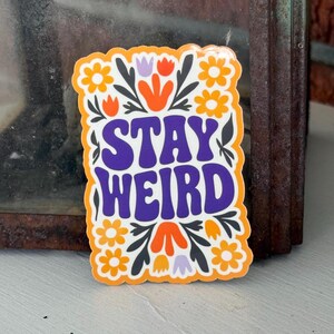 May include: A rectangular sticker with the words "STAY WEIRD" in purple, outlined in white, and set against a floral design. The sticker features orange and yellow flowers, green leaves, and orange and purple floral accents, all framed by an orange border.