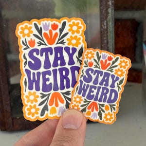 May include: Two stickers with the words "STAY WEIRD" in purple, surrounded by orange and white flowers, green leaves, and orange accents. The stickers have an orange border and a retro design.