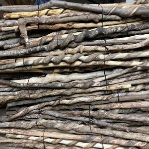 May include: A large collection of natural wooden walking sticks stacked horizontally and secured with black ties. Each stick features a unique, twisted spiral pattern carved into the bark, showcasing varying shades of brown, tan, and grey wood.