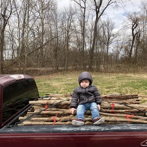 May include: A child wearing a dark gray hooded puffer jacket and blue jeans sits on a stack of brown wooden branches secured with red ties and black straps in the bed of a maroon pickup truck with a 4x4 emblem visible on the tailgate.