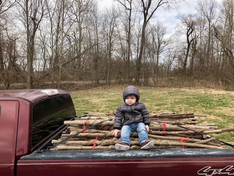 May include: A child wearing a dark gray hooded puffer jacket and blue jeans sits on a stack of brown wooden branches secured with red ties and black straps in the bed of a maroon pickup truck with a 4x4 emblem visible on the tailgate.
