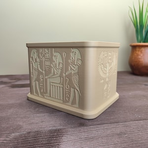 Può includere: Una scatola rettangolare beige con geroglifici e figure egizie in rilievo. La scatola presenta intagli dettagliati di faraoni, dei e simboli. Il design è di colore beige chiaro e la scatola poggia su una superficie di legno.