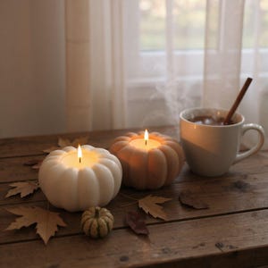 Op de afbeelding: Twee pompoenvormige kaarshouders met brandende kaarsen, één witte en één bruine, staan op een houten oppervlak. Een kleine kalebas en herfstbladeren zijn verspreid. Een witte mok met een kaneelstokje en een warm drankje staat op de achtergrond.