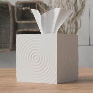 Puede incluir: Una caja de pañuelos blanca con un diseño texturizado de círculos concéntricos. La caja cuadrada está sobre una superficie de madera. Se ven pañuelos blancos saliendo de la parte superior. El fondo incluye cestas tejidas y elementos decorativos.