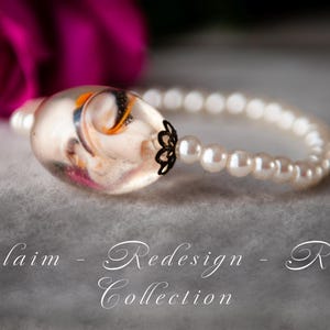 Pode incluir: Uma pulseira com uma conta oval transparente com desenhos embutidos, conectada a uma fileira de pérolas brancas. A conta tem uma tampa decorativa de metal preto. O texto "Reclaim - Redesign - Relove Collection" é visível.