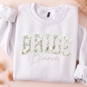 Könnte beinhalten: Ein weißes Sweatshirt mit Rundhalsausschnitt und dem Wort BRIDE in großen Blockbuchstaben, die mit einem weißen Blumenmuster und grünen Blättern gefüllt sind. Unter dem Wort BRIDE steht der Name Bianca in grauer Schreibschrift.