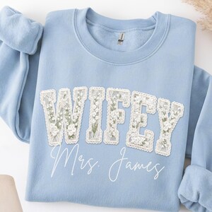 Könnte beinhalten: Ein hellblaues Sweatshirt mit Rundhalsausschnitt, das das Wort WIFEY in großen, weiß umrandeten Spitzenbuchstaben mit Blumenmuster zeigt. Darunter ist der Text Mrs. James in einer weißen, kursiven Schriftart aufgedruckt.