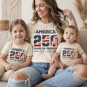 Könnte beinhalten: Eine Familie trägt passende cremefarbene Oberteile und einen Body mit dem Text "AMERICA 250 YEARS OF FREEDOM 1776-2026". Die Zahl 250 ist mit einem Sternen- und Streifenmuster gestaltet.