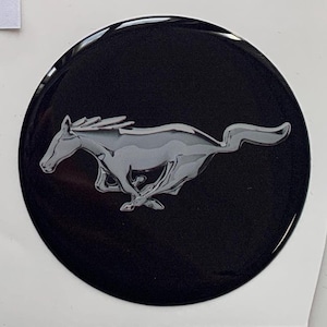 May include: A glossy black emblem featuring a silver, running horse silhouette. The horse is depicted in motion, with its legs extended. The emblem is circular and has a reflective surface, creating a sleek and modern look. The design is a classic automotive symbol.