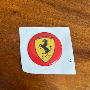 Peut inclure: Un autocollant rond avec le logo Ferrari. Le logo comprend un cheval cabré noir sur un écusson jaune, avec un bord vert, rouge et blanc. L'autocollant est sur un support blanc avec le numéro 42.