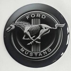 Peut inclure: Un emblème Ford Mustang noir et argenté. L'emblème comporte les mots "FORD" et "MUSTANG" en lettres blanches. L'image centrale est un cheval squelettique argenté en mouvement.