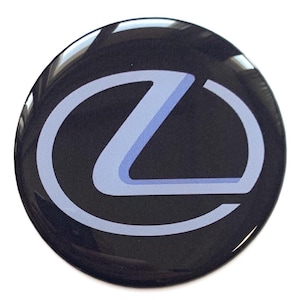Pode incluir: Um emblema circular e brilhante com o logotipo da Lexus. O logotipo é um 'L' estilizado dentro de um oval, com um esquema de cores azul claro e branco sobre um fundo preto. O emblema tem uma superfície reflexiva.