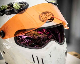 Może przedstawiać: Biały kask motocyklowy z pomarańczową osłoną, z logo "Bandit Helmets". Kask jest wykorzystywany jako donica, z małymi zielonymi roślinami i ziemią widocznymi wewnątrz i na górze.