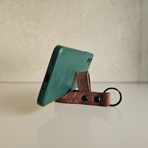 Puede incluir: Un teléfono inteligente verde azulado descansa sobre un soporte de teléfono de cuero marrón. El soporte está hecho de una pieza de cuero curvada con un lazo y un llavero. El soporte tiene las letras "ARTM" estampadas.