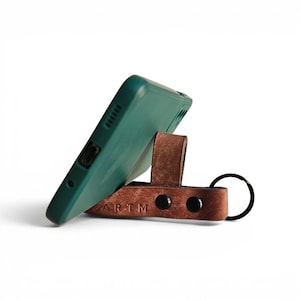 May include: A dark green smartphone case propped up by a brown leather strap with a black metal key ring. The leather strap features two black metal snaps and the embossed text ARTM, functioning as a stand for the mobile device.