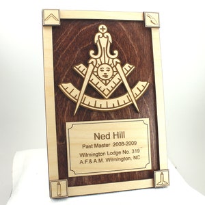 May include: A wooden plaque with a brown background and a light wood trim. The plaque features a Masonic symbol with a compass and square, and a text plaque that reads "Ned Hill Past Master 2008-2009 Wilmington Lodge No. 319 A.F.& A.M. Wilmington, NC".