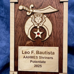 May include: A wooden plaque with a brown background and a light wood frame. The plaque features a crescent moon and star design with a red jewel in the centre. The text "Leo F. Bautista AAHMES Shriners Potentate 2025" is engraved on the plaque.