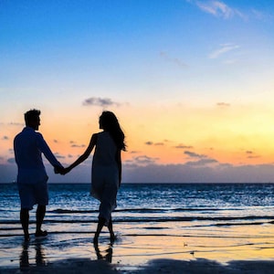 May include: Silhouette of a couple holding hands while walking on a beach at sunset. The sky transitions from blue to orange and yellow, reflecting on the wet sand and ocean. The couple is dressed in light-colored clothing.
