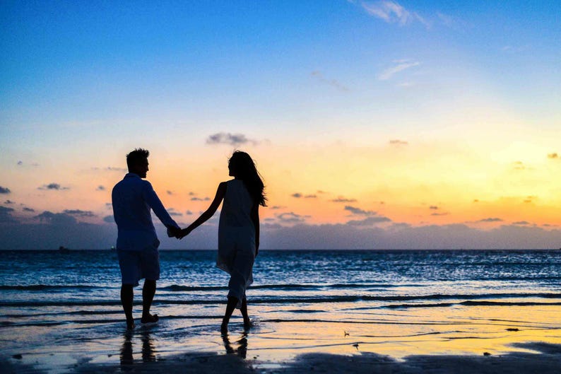 May include: Silhouette of a couple holding hands while walking on a beach at sunset. The sky transitions from blue to orange and yellow, reflecting on the wet sand and ocean. The couple is dressed in light-colored clothing.