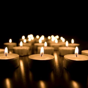 May include: A collection of lit tea light candles against a dark background. The small, round candles are arranged in a group, with their flames casting a warm, golden glow. The scene evokes a sense of tranquility.