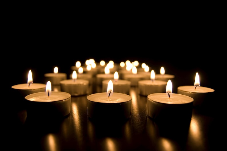 May include: A collection of lit tea light candles against a dark background. The small, round candles are arranged in a group, with their flames casting a warm, golden glow. The scene evokes a sense of tranquility.