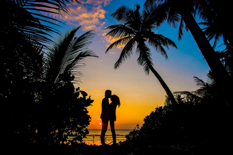 May include: Silhouette of a couple kissing at sunset on a beach. Palm trees frame the scene against a vibrant sky of orange, yellow, and blue hues. The sun is setting over the ocean.