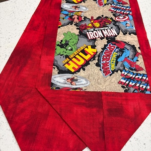 Può includere: Una tovaglia rossa e multicolore con personaggi dei fumetti. Il pannello centrale mostra immagini di The Hulk, Iron Man e Spider-Man. I bordi sono in tessuto rosso.