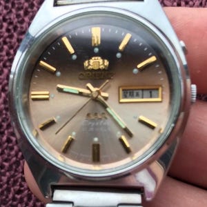 May include: A vintage Orient watch with a silver-tone stainless steel band and case. The watch face has a gradient brown color with gold-tone hour markers and hands. The watch displays the date and day.