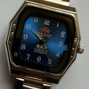 May include: A silver-toned Orient wristwatch with a blue dial. The watch has a rectangular face with a black border and a silver-toned metal band. The dial displays the Orient logo, numerals, and the text "23 Jewels Automatic."