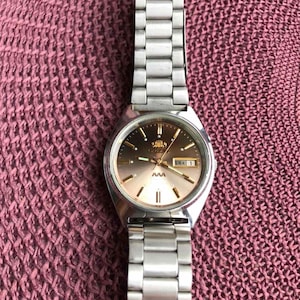 Puede incluir: Un reloj de pulsera plateado con una esfera degradada marrón y dorada y una correa de eslabones plateada. La esfera del reloj muestra el día y la fecha. El reloj está sobre una superficie rosa texturizada.
