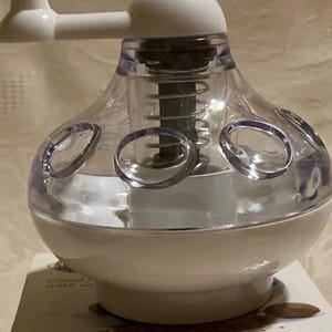 Puede incluir: Un molinillo de especias blanco y transparente con un asa y una parte superior redonda. El molinillo tiene un cuerpo transparente y bulboso con aberturas circulares. El texto "NORPRO Spice Grinder" es visible en la caja de abajo.