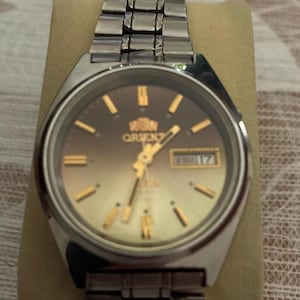 May include: A silver-toned Orient watch with a brown and gold gradient dial. The watch features gold-colored hour markers and hands, and a day-date display. The watch has a stainless steel band.