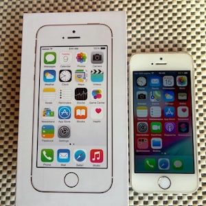 Puede incluir: Un teléfono inteligente blanco con un botón de inicio y una pantalla con iconos de aplicaciones coloridos, colocado junto a su caja de embalaje rectangular blanca original que muestra una imagen impresa del mismo modelo.