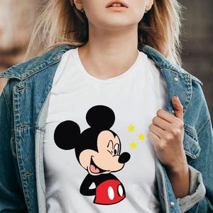 May include: White t-shirt featuring a smiling Mickey Mouse graphic with black ears, a red shorts, and yellow stars. The shirt is worn with a blue denim jacket.